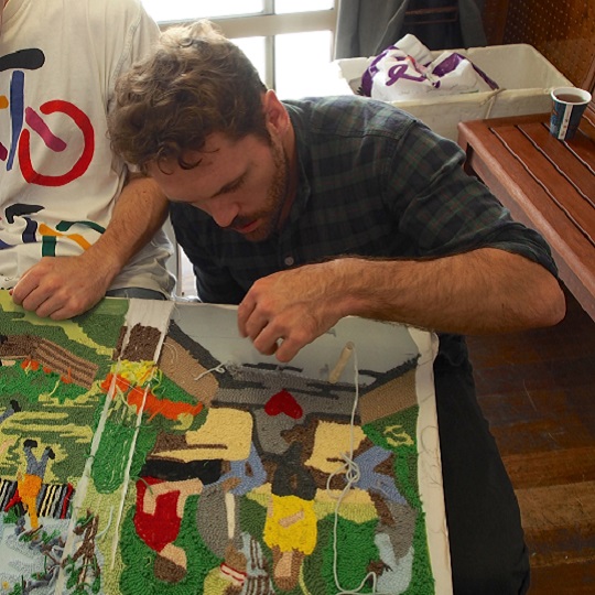 Social designer Tasman Munro crouches at side of table with tapestry needle in hand working on a tapestry, colourful wool soft fabric